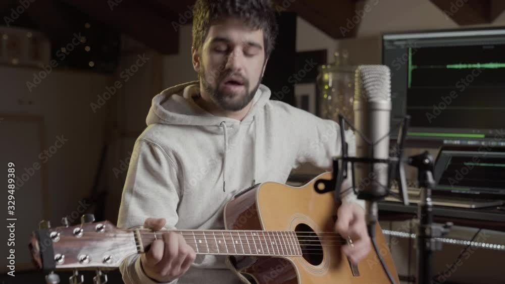 Attractive creative male vocalist in real home recording studio, playing guitar in front of condenser microphone, audio wave form on computer monitor screen in audio software - parallax motion shot.