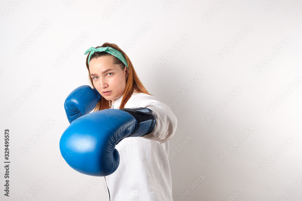 Girl power concept. Confident young woman isolated on a gray wall background. Female and independent power. The girl raises her hand in a boxing glove to strike.Copy space. Girl power concept. Confident young woman isolated on a gray wall background. Female and independent power. The girl raises her hand in a boxing glove to strike.Copy space.