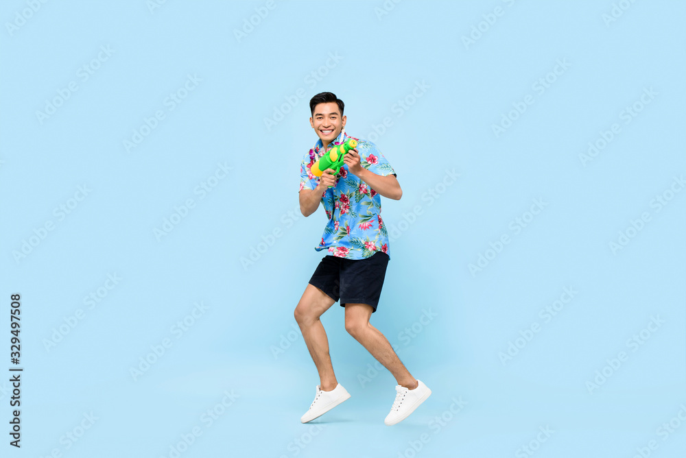Handsome smiling young Asian man playing with water gun and jumping in studio blue background for Songkran festival in Thailand and southeast Asia Handsome smiling young Asian man playing with water gun and jumping in studio blue background for Songkran festival in Thailand and southeast Asia