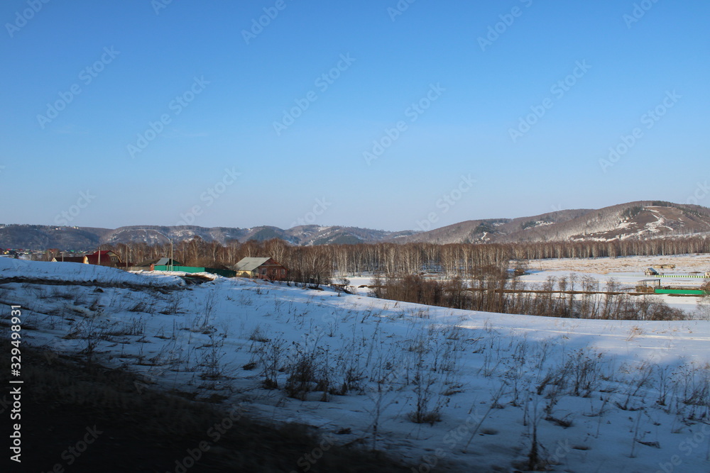 Rural houses on the background of snow covered fields trees and mountains under a blue sky.