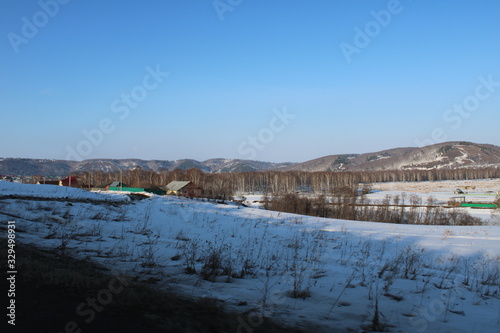 Rural houses on the background of snow covered fields trees and mountains under a blue sky.