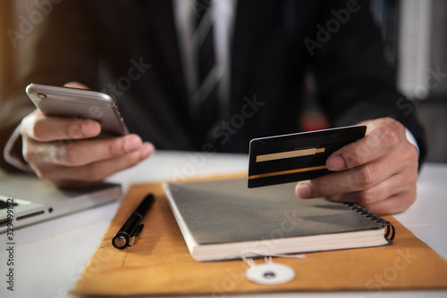 Male businessman use credit cards to conduct financial transactions through phones, tablet, and laptop in office.