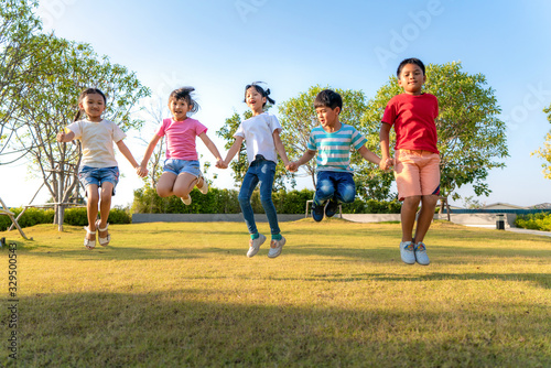 Large group of happy Asian smiling kindergarten kids friends holding hands playing and jumping together during a sunny day in casual clothes at city park.