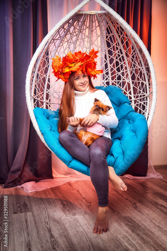Beautiful brunette girl whth long hair in the blue shirt, with a wreath of yellow autumn maple leaves and small dog on a white wicker hanging chair with pillow photo