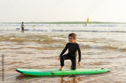 Little boy standing on surfboard. Adorable child in wetsuit standing on surfboard and looking aside on ocean coast. Surfing concept