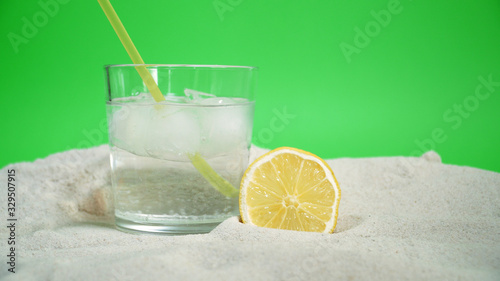 glass with ice and soda water and lemon on the sand on a green background