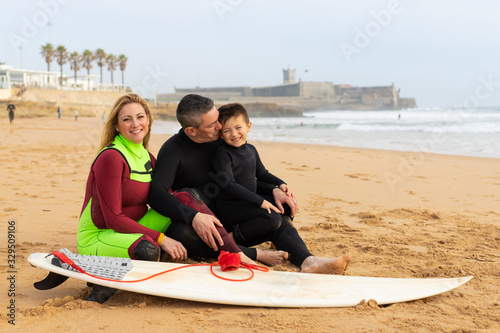 Happy family with surfboard on ocean coast. Cheerful parents and cute little son in wetsuits sitting together on sandy coast. Water sport concept