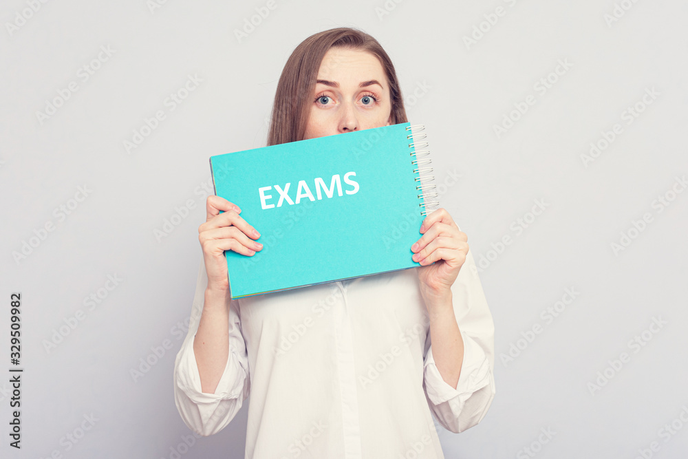 Girl hides behind a blue notebook with inscription: exams, grey background, copy space Girl hides behind a blue notebook with inscription: exams, grey background, copy space