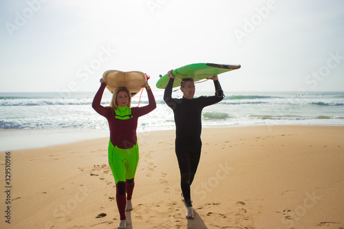 Couple with surfboards walking on beach. Front view of middle aged man and woman in wetsuits holding surfboards and walking on sea coast. Surfing concept