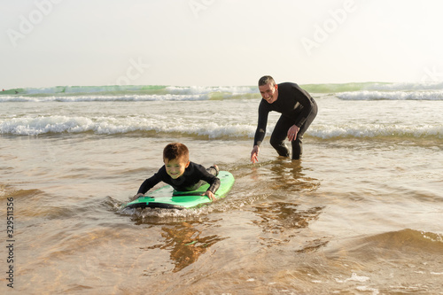 Father teaching son surfing in ocean. Happy father helping little son in wetsuit lying and swimming on surfboard on waves. Surfing concept