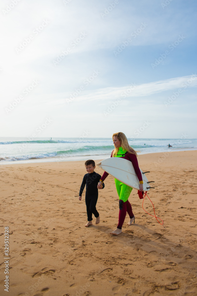 Mother and son in wetsuits walking on beach. Cheerful mother with surfboard and cute little son holding hands and walking on sea coast. Surfing concept