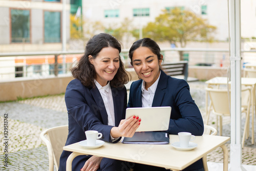 Content businesswomen with tablet pc in outdoor cafe. Cheerful female colleagues sitting at table, drinking coffee and using digital tablet in outdoor cafe. Wireless technology concept