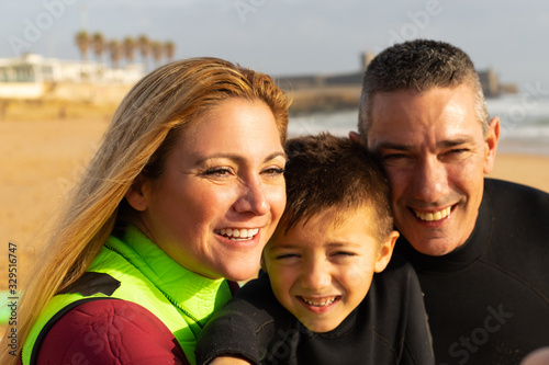 Happy family in wetsuits on beach. Close-up portrait of cheerful middle aged parents and cute little son in wetsuits smiling at camera on sea coast. Water sport concept