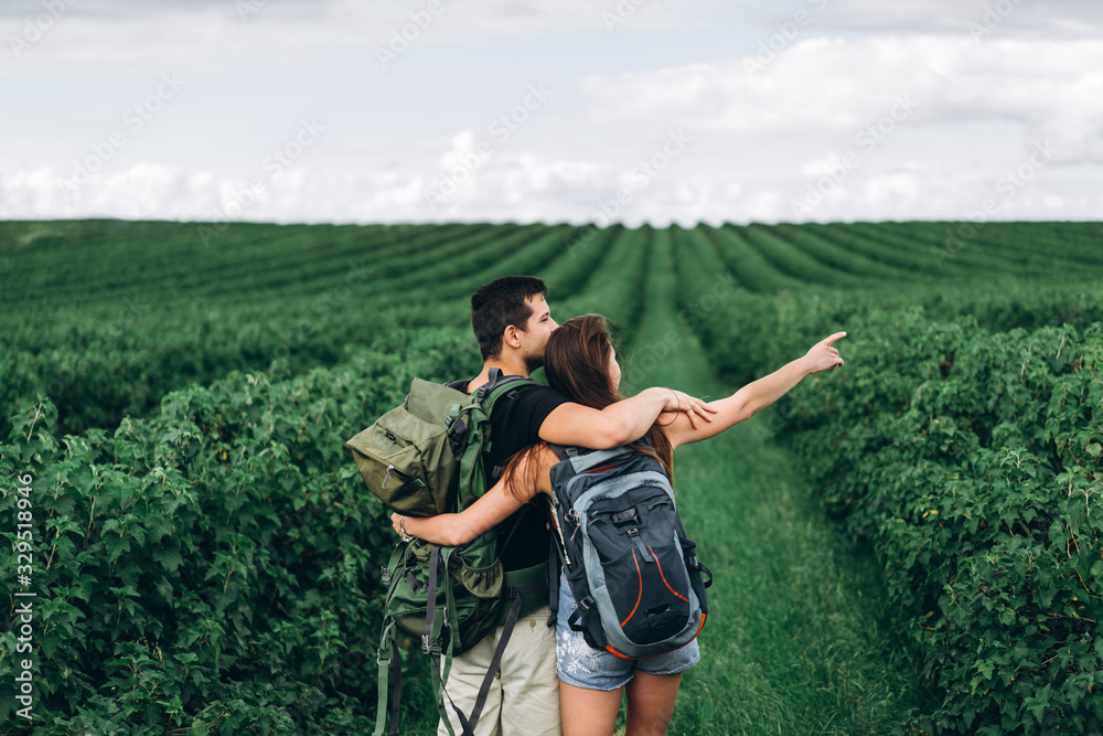 Back view of young couple with backpacks on currant plantations. Woman with long hair points her hand to the horizon