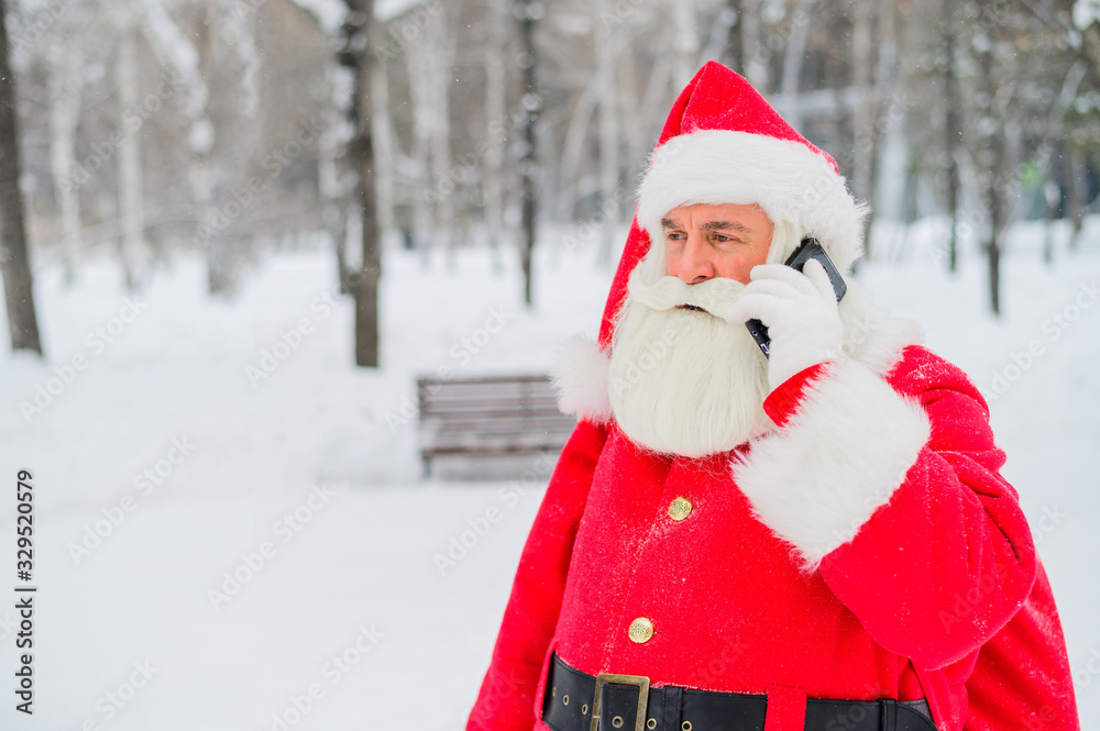 Surprised santa claus is using mobile outside. Santa Claus stands in the background of buildings in winter and calls on a smartphone. Snow in December. Surprised santa claus is using mobile outside. Santa Claus stands in the background of buildings in winter and calls on a smartphone. Snow in December.
