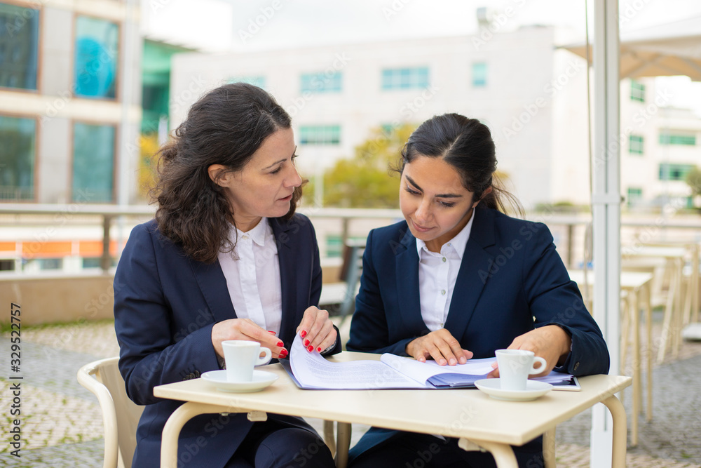 Businesswomen working with papers in outdoor cafe. Professional female colleagues in formal wear sitting at table, drinking coffee and discussing work in outdoor cafe. Coffee time concept Businesswomen working with papers in outdoor cafe. Professional female colleagues in formal wear sitting at table, drinking coffee and discussing work in outdoor cafe. Coffee time concept