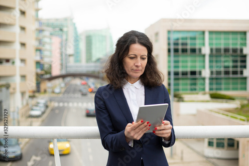 Focused woman using tablet pc. Serious middle aged businesswoman using digital tablet while standing on urban city street. Wireless technology concept © Mangostar