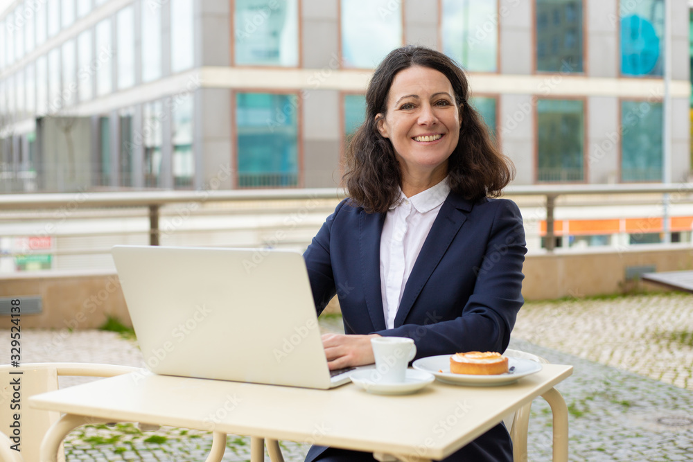 Businesswoman using laptop and smiling at camera. Happy middle aged businesswoman working with laptop computer while sitting at table with coffee outdoors. Technology concept Businesswoman using laptop and smiling at camera. Happy middle aged businesswoman working with laptop computer while sitting at table with coffee outdoors. Technology concept