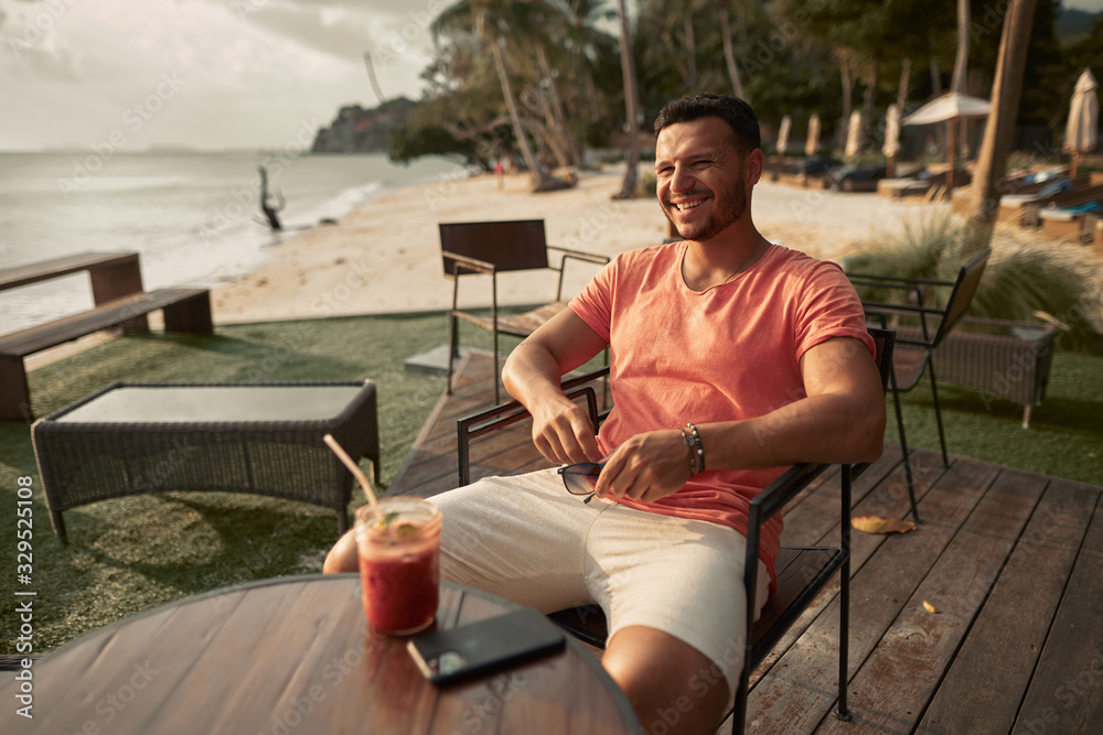 A handsome man with a fruit cocktail sits at a table at the beach bar, enjoying the sunset