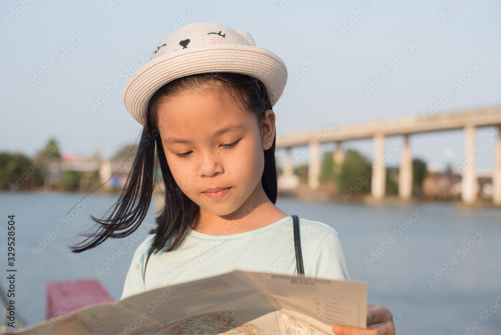 asia little girl with city map on summer vacation. travel and adventure concept, outdoor portrait girl  in hat  traveling around new place and searching beautiful sights. town by the river.