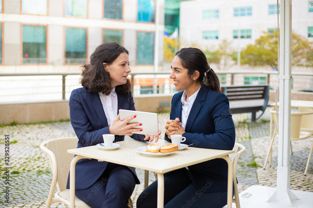 Businesswomen drinking coffee and using tablet pc. Professional female coworkers in formal wear sitting at table and drinking coffee together in outdoor cafe. Coffee time concept