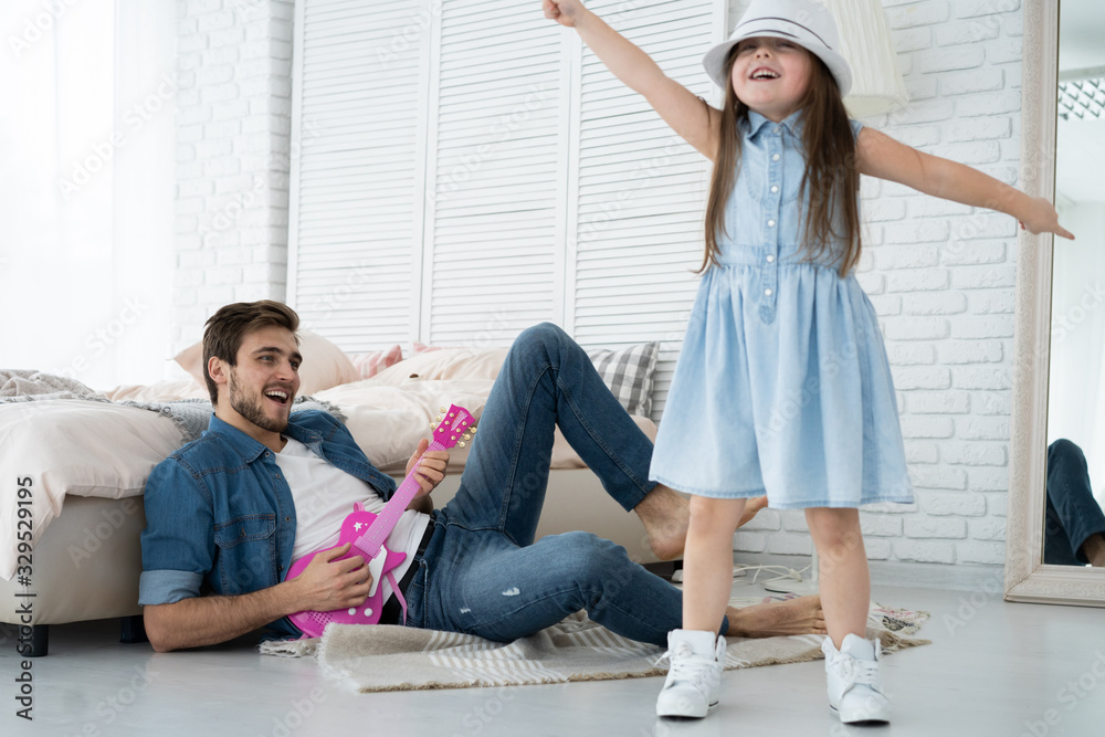 Having fun together. Young father playing guitar for his little daughter and smiling while spending free time at home.