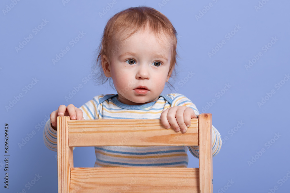 Close up portrait of little child looking aside, standing on chair isolated over blue background in studio, wearing striped sweater, being attentive, focused on something. Childhood concept. Close up portrait of little child looking aside, standing on chair isolated over blue background in studio, wearing striped sweater, being attentive, focused on something. Childhood concept.