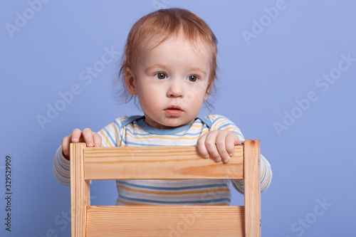 Close up portrait of little child looking aside, standing on chair isolated over blue background in studio, wearing striped sweater, being attentive, focused on something. Childhood concept.