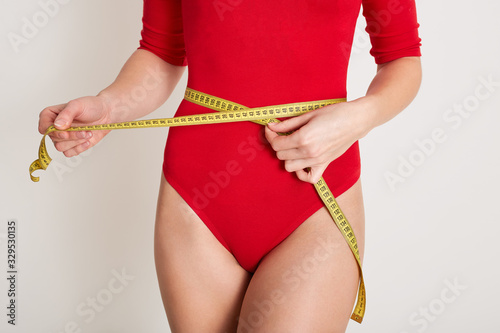 Indoor picture of unknown female body measuring her waist with yellow measuring tape, caring about her body, keeping fit, losing weight, being on diet, wearing red combidress. Women and diet concept.
