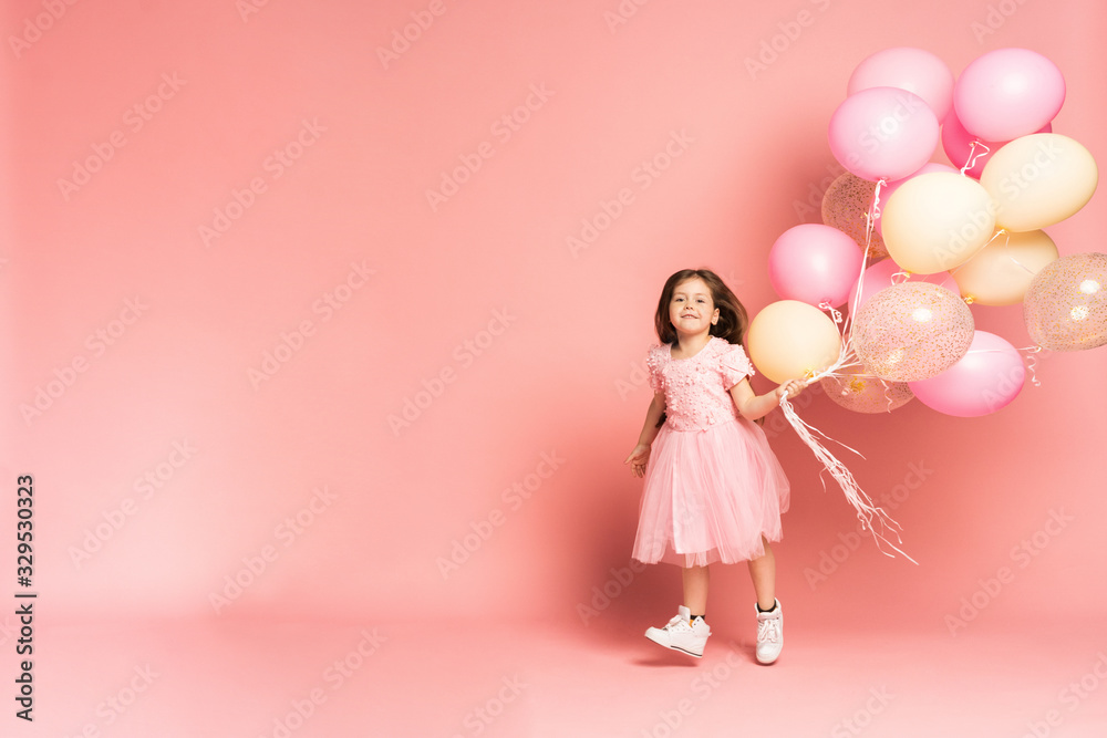 Happy celebration of birthday party with flying balloons of charming cute little girl in tulle dress smiling to camera isolated on pink background. Charming smile, expressing happiness