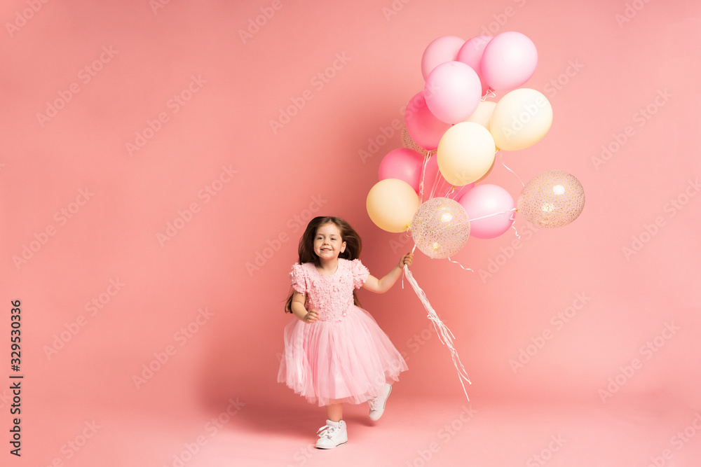 Happy celebration of birthday party with flying balloons of charming cute little girl in tulle dress smiling to camera isolated on pink background. Charming smile, expressing happiness