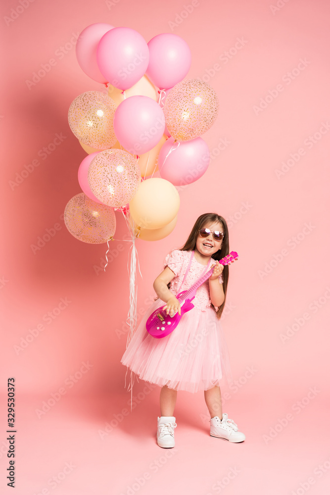 Laughing fashion little girl having fun with guitar isolated on pink background.