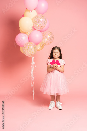 Happy celebration of birthday party with flying balloons of charming cute little girl in tulle dress holding red paper heart and smiling to camera isolated on pink background