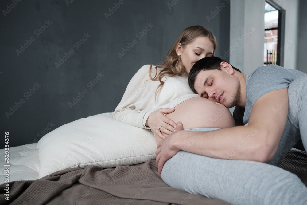 Handsome man and his beautiful pregnant wife are hugging and smiling sitting at the sofa at home. Handsome man and his beautiful pregnant wife are hugging and smiling sitting at the sofa at home.