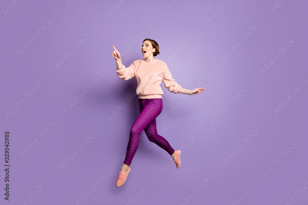 Full length photo of funny lady jumping up high see low shopping prices direct finger advert banner walking down street wear casual stylish pullover pants isolated purple color background