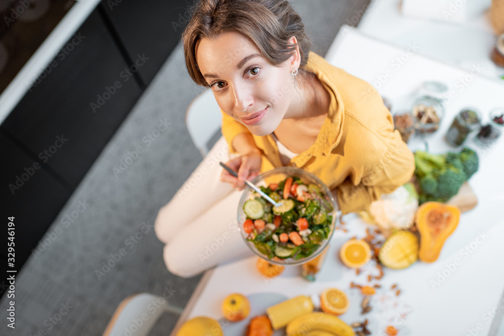 Portrait of a young cheerful woman eating salad at the table full of healthy raw vegetables and fruits on the kitchen at home, view from above. Concept of vegetarianism, healthy eating and wellness Portrait of a young cheerful woman eating salad at the table full of healthy raw vegetables and fruits on the kitchen at home, view from above. Concept of vegetarianism, healthy eating and wellness
