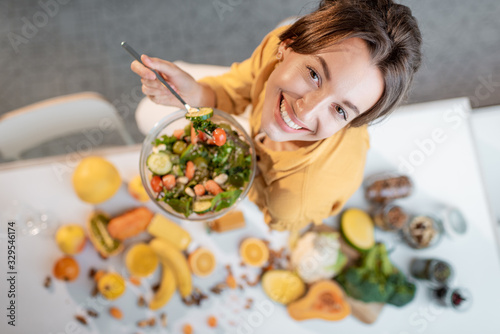 Fotografia Portrait of a young cheerful woman eating salad at the table full of healthy raw vegetables and fruits on the kitchen at home, view from above
