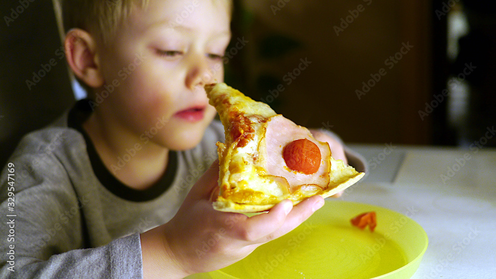 Dziecko zjada kawałek pizzy. Chłopiec trzyma pizzę i patrzy na nią, jest głodny i zjada posiłek ze smakiem. Dzieci niejadki zastanawiają się nad tym czy zjeść. - obrazy, fototapety, plakaty 