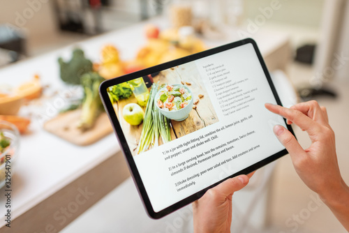 Woman looking on the digital recipe, using touchscreen tablet while cooking healthy meal on the kitchen at home, close-up view on the screen photo