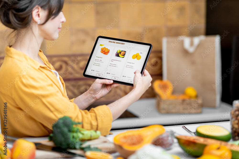 © rh2010 - Woman holding digital tablet with launched online shopping market while standing on the kitchen with lots of fresh food on the table