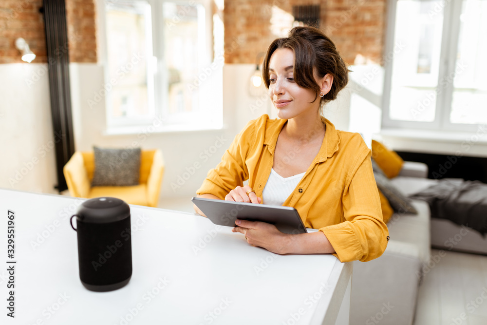 Happy young woman controlling smart home devices with a voice commands and smart speaker at home. Concept of a smart home and managing wireless devices remotely Happy young woman controlling smart home devices with a voice commands and smart speaker at home. Concept of a smart home and managing wireless devices remotely