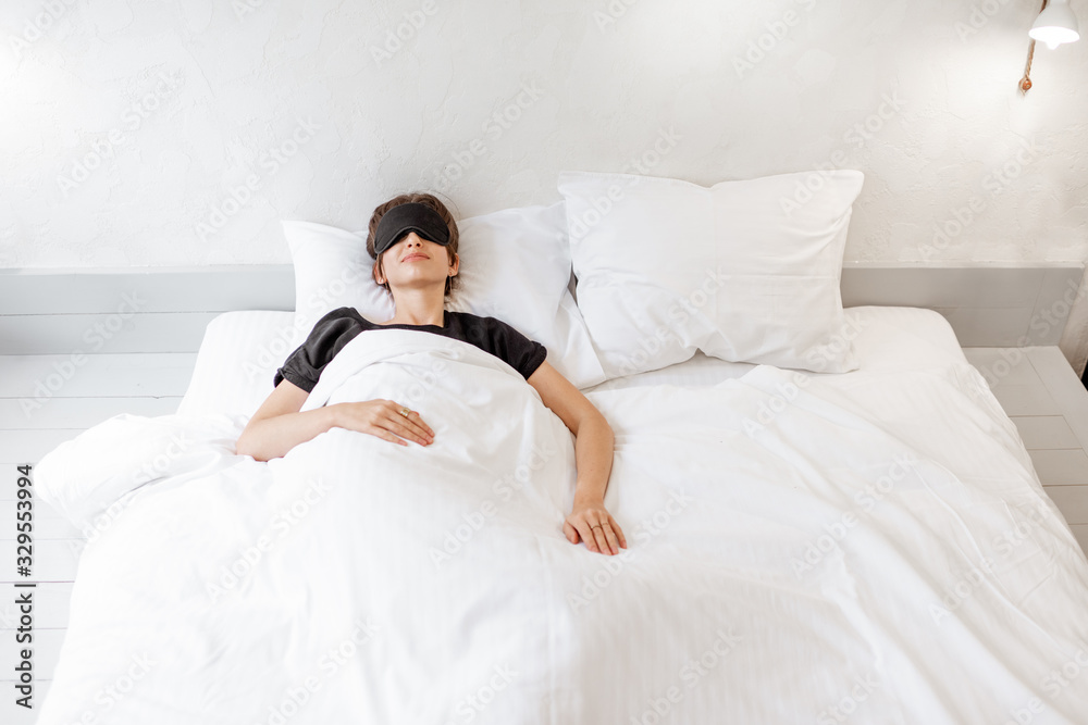 Woman lying with a sleeping mask in bed covered with white sheets in bright bedroom in the morning Woman lying with a sleeping mask in bed covered with white sheets in bright bedroom in the morning