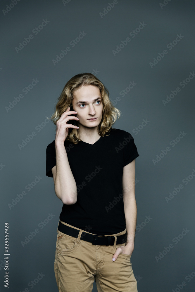 Young handsome guy with long blonde hair is talking on the phone on a gray background.