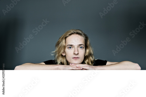 A young attractive man peeks out from above a white wall on a gray background. Looks surprised laid his head in his hands. The elbows are apart.