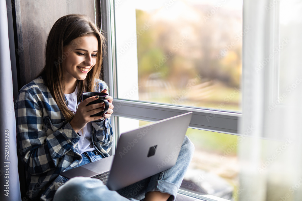 Young woman with coffee cup and laptop sitting on widnowsill at home
