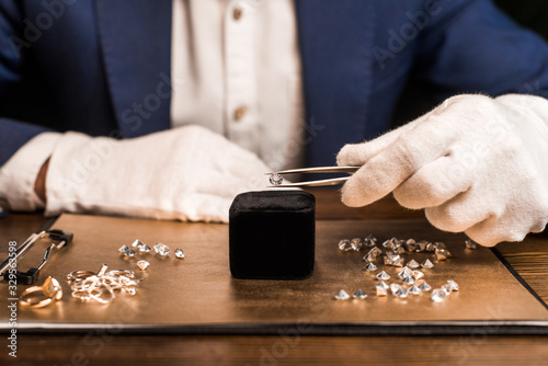 Cropped view of jewelry appraiser holding gemstone in tweezers near jewelry on board on table isolated on black photo