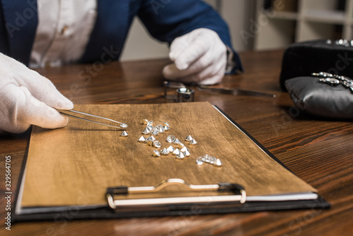 Cropped view of jewelry appraiser with pliers examining gemstones on wooden board on table in workshop