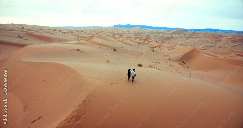 Young couple walking together on sandy hills endless desert Aerial following shot of a young couple walking on sands over hills of desert long shot Young couple walking together on sandy hills endless desert Aerial following shot of a young couple walking on sands over hills of desert long shot