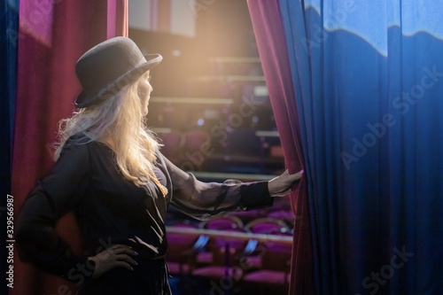 A blonde caucasian girl in black overalls and a bowler hat looks from the curtains in the theater hall.  The concept of the show must begin. Back view.
