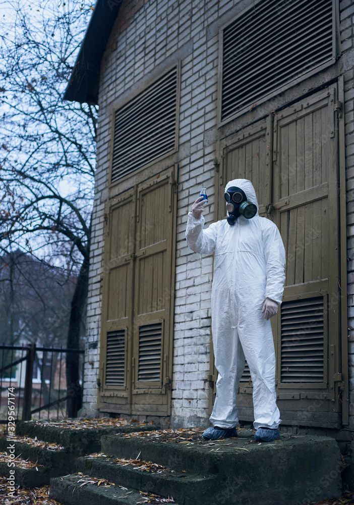 Vertical full length snapshot of a scientist standing in front of an old door and observing reaction in glass flask with blue liquid. Ecologist wearing white uniform, gloves and gas mask